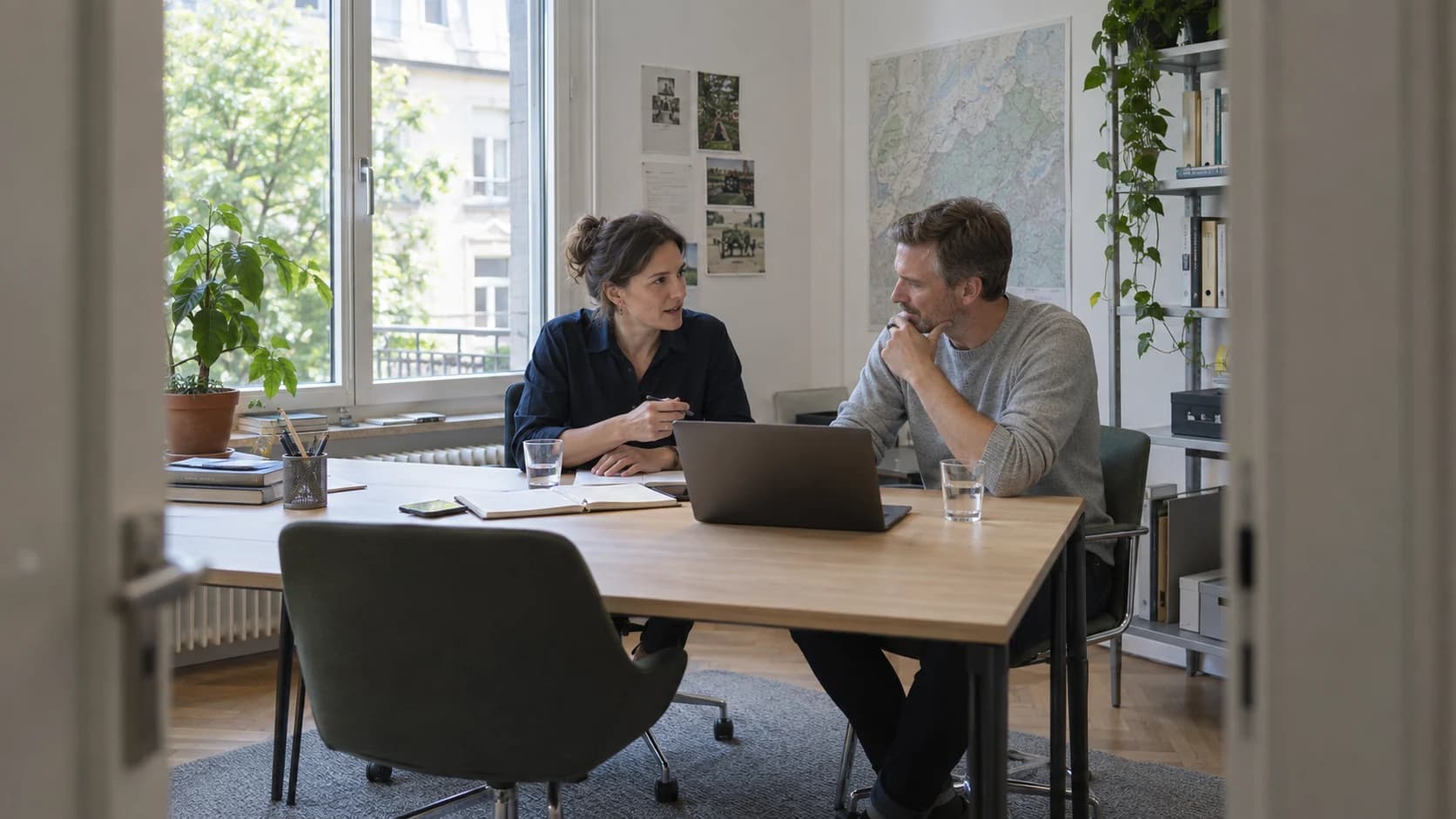 Zwei Kollegen besprechen ihre Arbeit an einem Eichentisch in einem Büro mit natürlichem Licht.